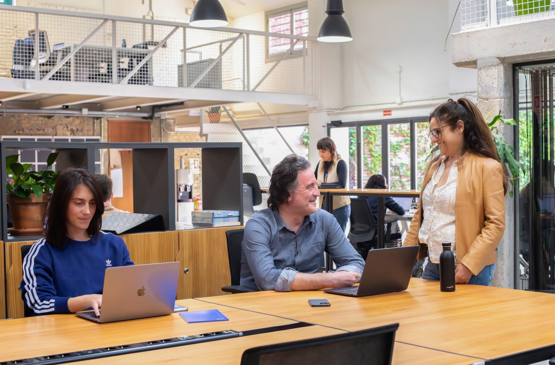 Una mujer joven sentada en una mesa compartida en un coworking junto a un hombre más mayor, los dos con portátil, intractuando con una mujer de mediana edad que está de pie hablando con ellos sonriendo.
