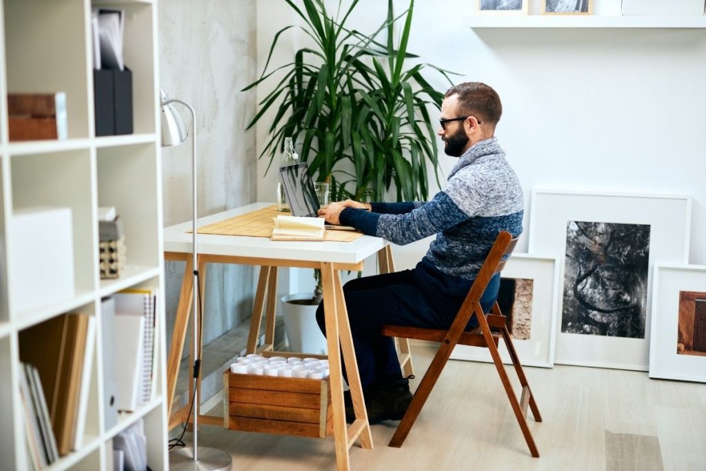 Hoobre sentado en mesa de escritorio blanca con una panta al fondo y una estaneria con libros y cuadros apoyados en el suelo.