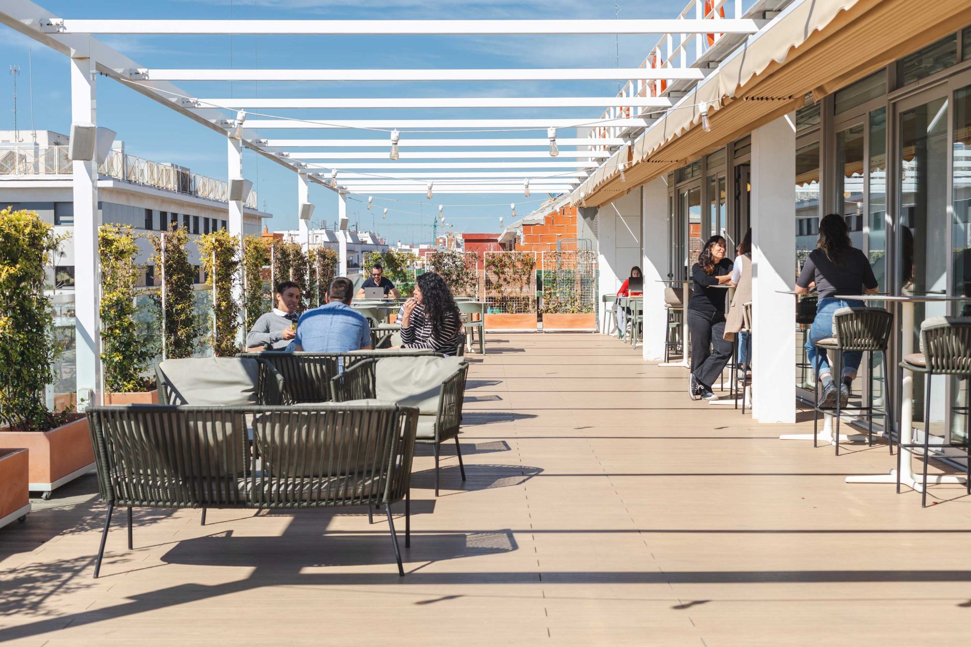 Imagen de terraza en un ático de edificio soleada con varias personas en distintas mesas charlando o trabajando con su portátil