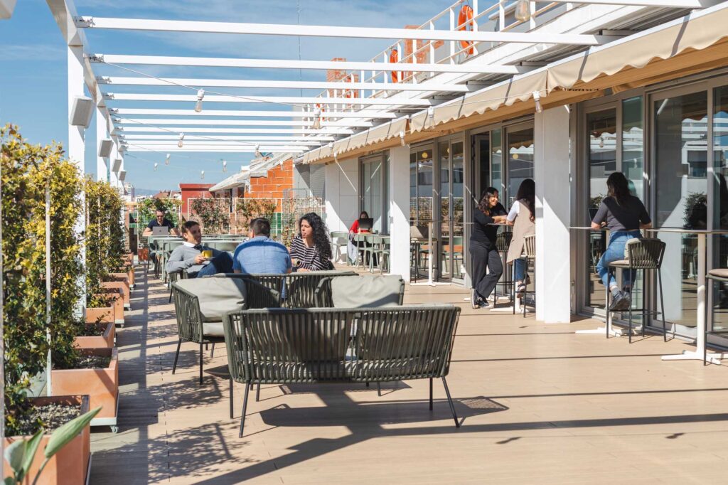 Imagen de una terraza en el ático de un edificio con sol y gente sentada en mesas altas y bajas con sus portátiles o tomando café