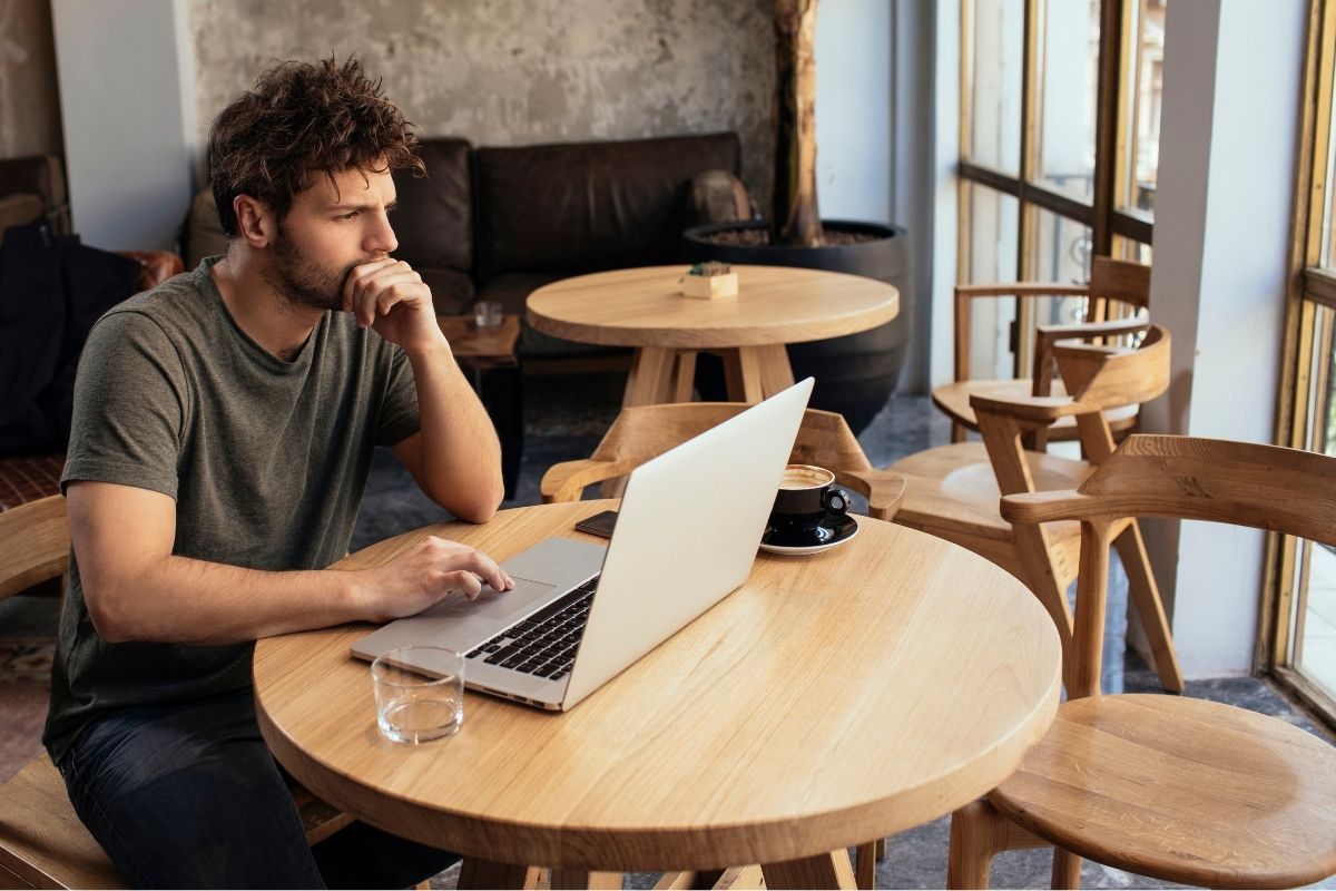 Un hombre joven sentado a una mesa redonda de una cafetería coworking con un café y un portátil.